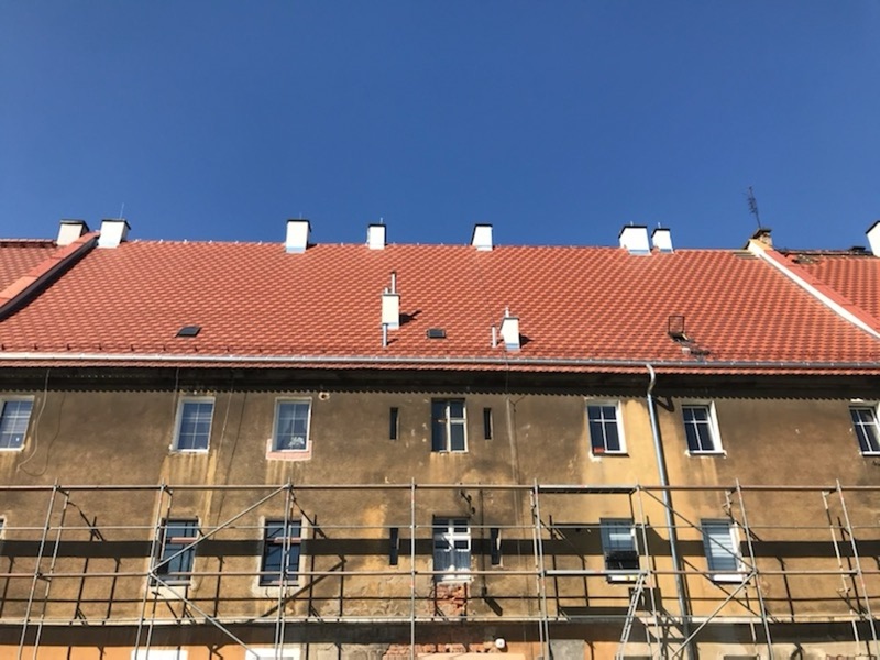 Kamienica z nowym pokryciem z czerwonej dachówki i białymi kominami, przy elewacji rusztowanie