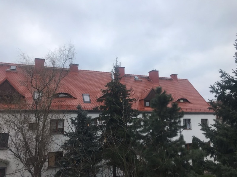 Dach z czerwonej dachówki z lukarnami i oknami dachowymi, częściowo zasłonięty drzewami